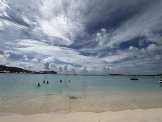 A wide Caribbean beach with calm, clear water and a few swimmers under dramatic streaked clouds