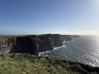 The Cliffs of Moher dropping into a sunlit Atlantic Ocean, seen from the grassy clifftop in Ireland