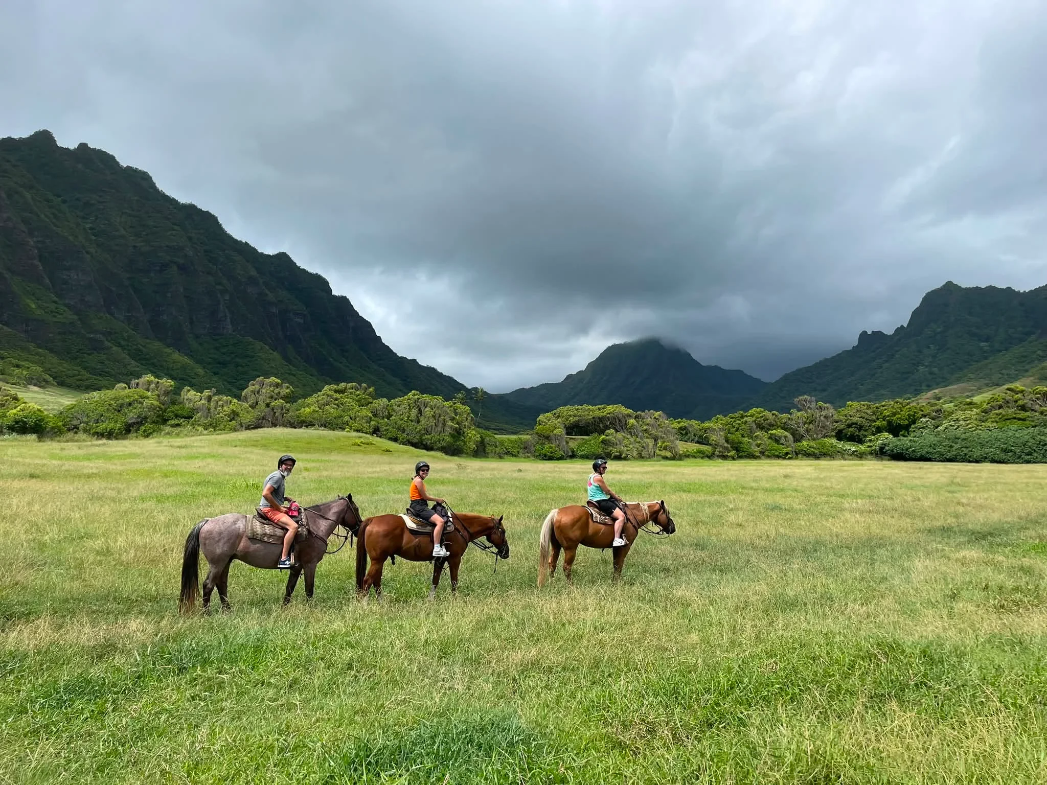 Three horseback riders crossing a green valley beneath steep cloud-covered mountains in Hawaii