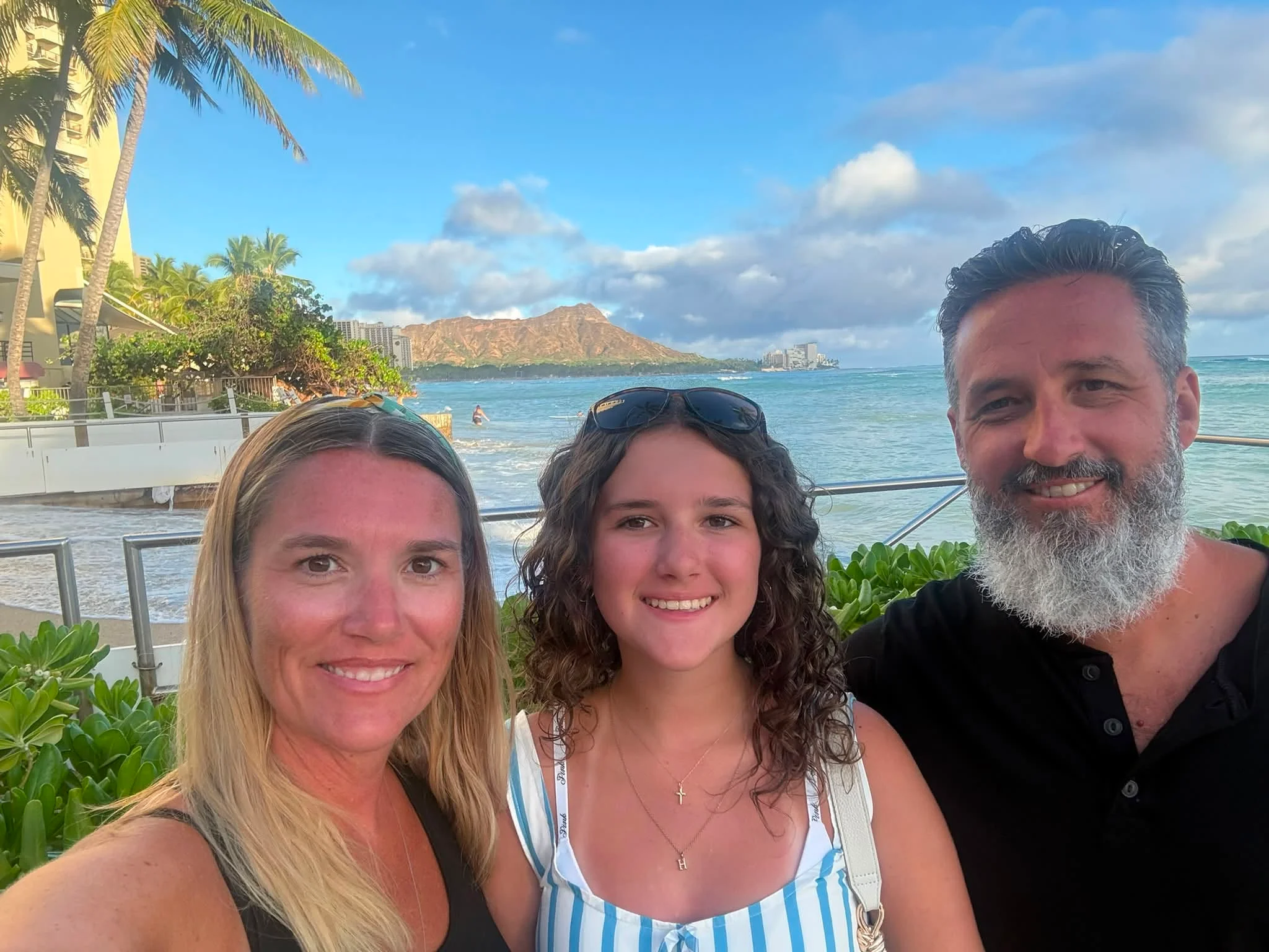 A family of three taking a selfie on the Waikiki shoreline with Diamond Head crater behind them
