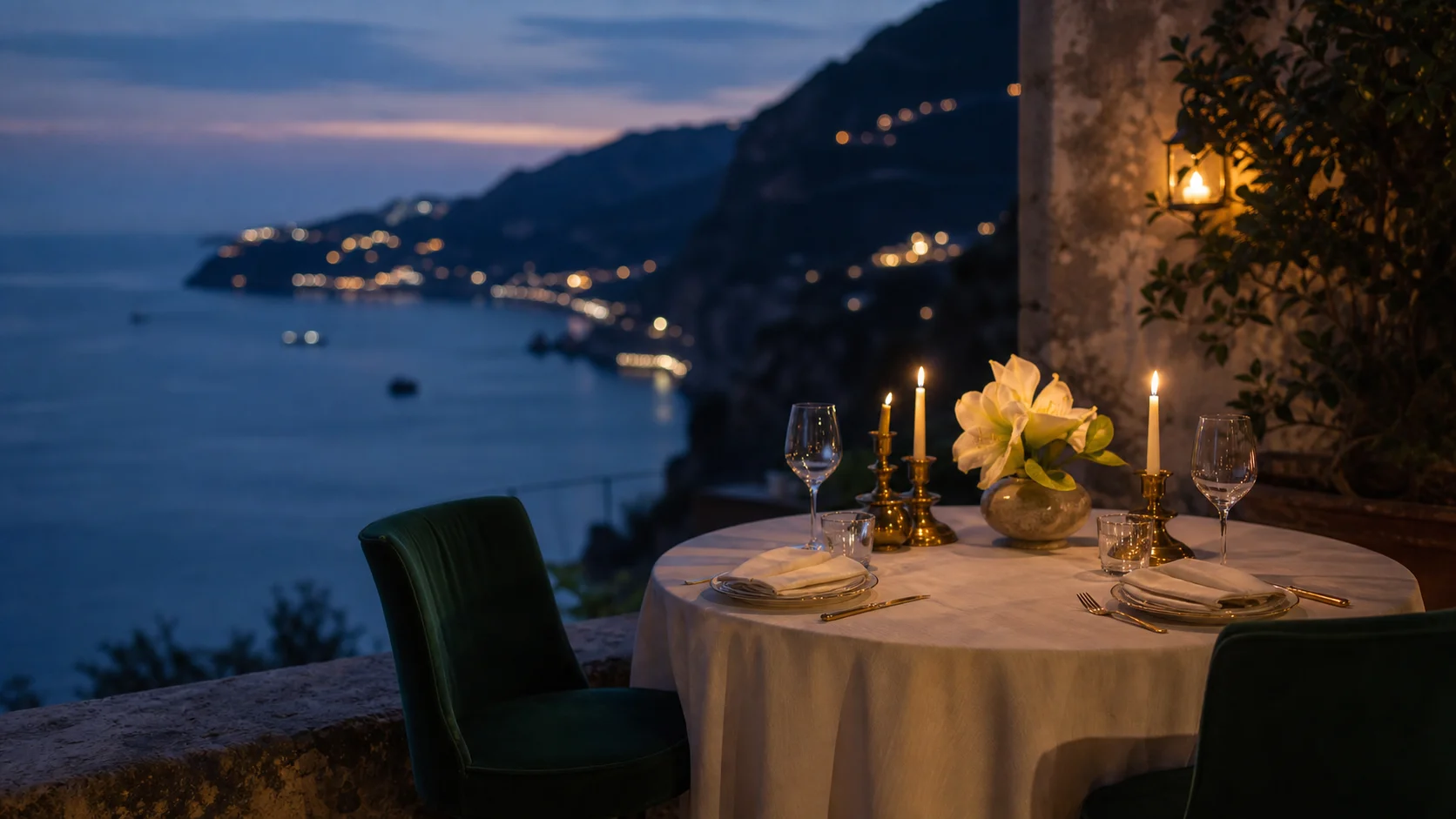 A candlelit dinner table for two on a stone terrace at dusk, overlooking coastal cliffs and town lights